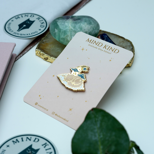 A gold and blue enamel pin shaped like a crescent moon and flower, mounted on a light pink card labeled 'MIND KIND'. The card is styled on a white surface with crystals, a notebook, and a green leaf.