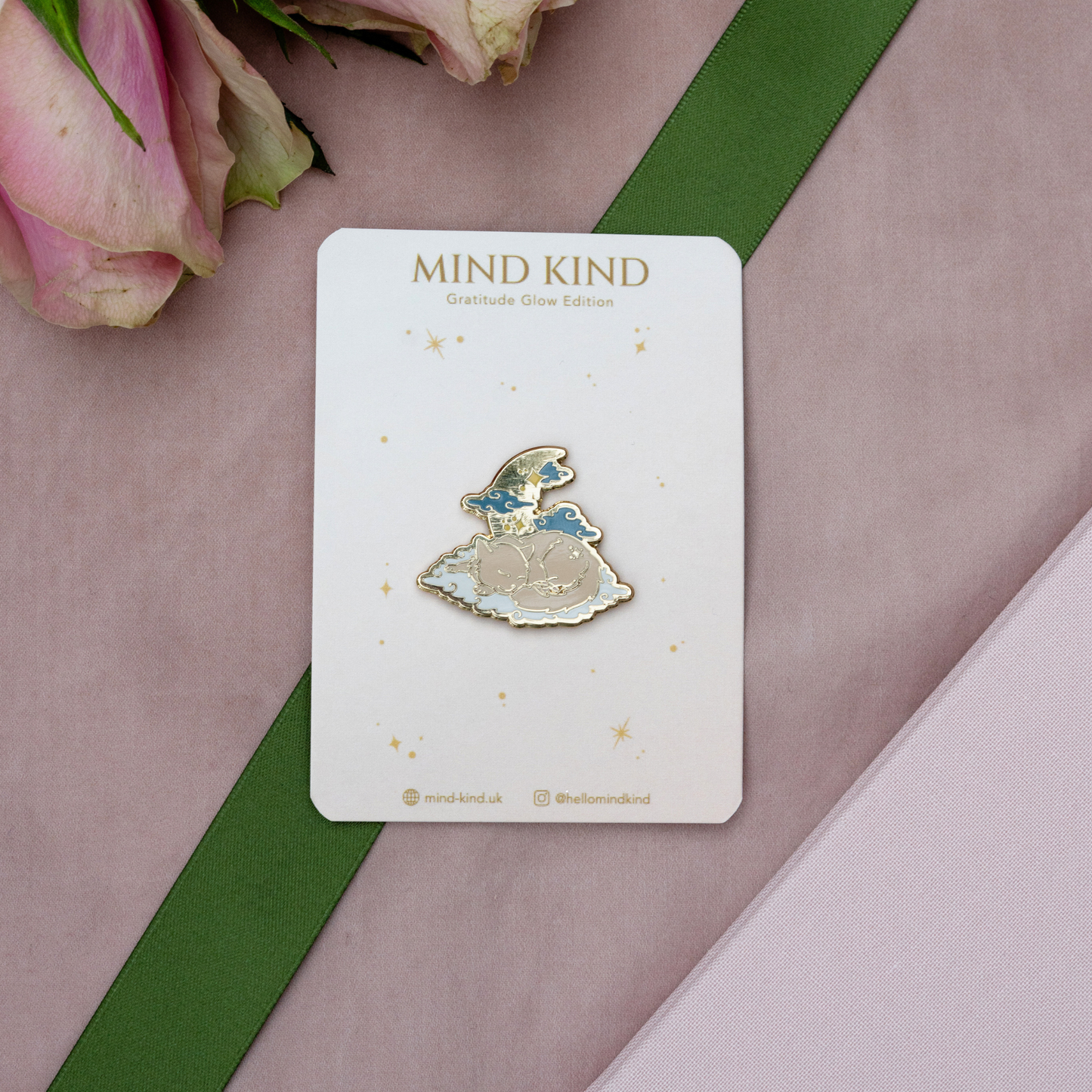 A small enamel pin of a fairy in a dress on a white card, labeled 'MIND KIND Gratitude Glow Edition,' styled on a pink surface with a green ribbon and pink flowers.